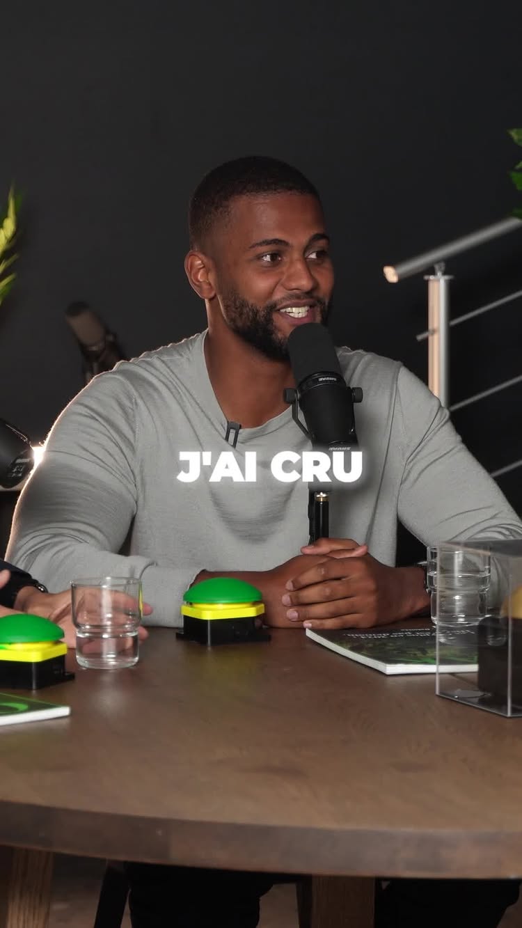 A man is seated at a table, speaking into a microphone. The text overlayed reads "J'AI CRÛ" in a bold typeface. Behind him, there's a dark background, and two microphones are visible. The table is composed of a dark wooden material, and there are two green and yellow buttons and water glasses on the table.