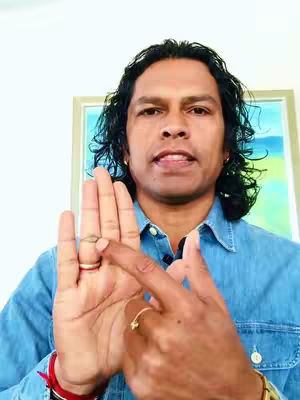 A medium shot of a man with dark curly hair and a blue denim shirt. He is holding up his left hand, palm facing the viewer, with the index finger of his right hand extended and pointing towards the ring finger on the left hand. Behind him is a blurry, framed picture on a light-colored wall. The background is a plain, bright wall