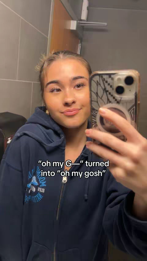 A young woman is taking a mirror selfie in a bathroom. She is wearing a dark blue hoodie and holding her phone up to take the picture. The bathroom has light gray tiled walls, and a mirror reflection shows a gray towel dispenser and a partially obscured door. Text overlay on the image states "oh my G-- turned into 'oh my gosh.'"