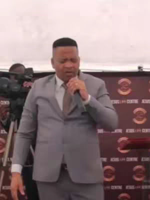A man in a gray suit stands in front of a blurred background, holding a microphone. He is speaking, and the background has a repeating logo pattern.