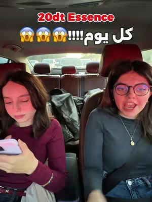 Two young women are in a car. The woman on the left is looking down at her phone while the woman on the right is looking at the camera with her mouth open. Text in a red box at the top of the image says "20dt Essence" in red and white Arabic script. There is text in white in Arabic script. The background of the car is visible.