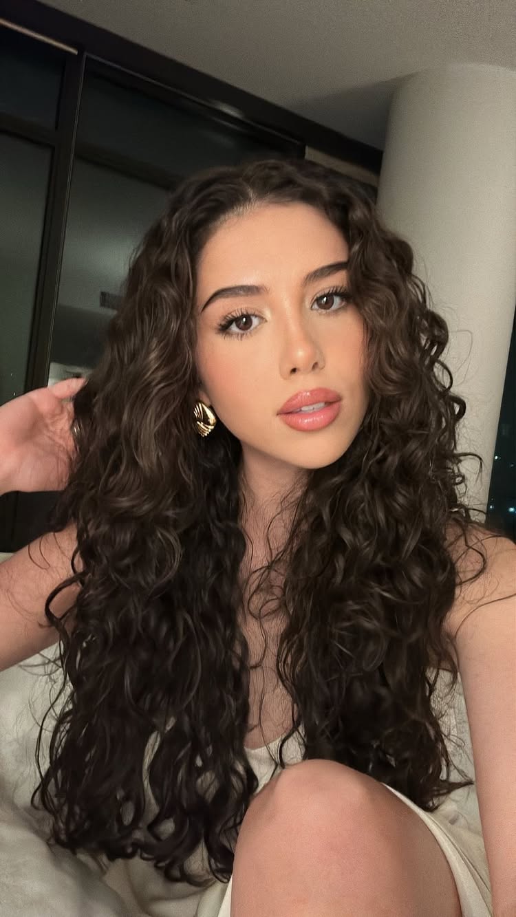 The cover image is a selfie of a young woman with long, dark curly hair. She is looking directly at the camera. She's wearing gold hoop earrings and has light makeup, with a focus on her lips. The background suggests an indoor setting, possibly a room in a building at nighttime with a window. The lighting is soft, and the composition is a close-up, focusing on the woman's face and upper body.