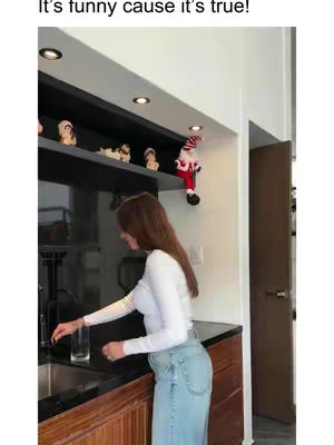 A woman in a kitchen setting, reaching for a glass while standing in front of a counter. The background includes overhead cabinets with decorative items and a decorative shelf. The lighting is bright, highlighting the subjects in the image. The woman is in a white top and blue jeans.