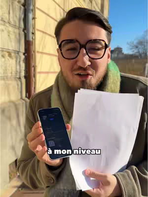 A man wearing glasses and a scarf is holding a smartphone and a stack of white papers. The background features a building facade and a clear sky.