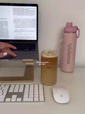 A well-lit, top-down shot on a white surface features a neatly organized study setup. A laptop displays text, with a water bottle and a glass of iced coffee in the background. A white keyboard and mouse are in the frame. The text overlay reads "morning routine".
