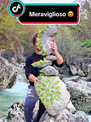 A man stands outdoors near a flowing stream, balancing a stack of large, irregular stones decorated with green leaves. The stones are arranged to resemble a sculpture. Above, a text overlay reads 'Meraviglioso' in a white font with a black outline against a color-blocked background with the Tiktok logo
