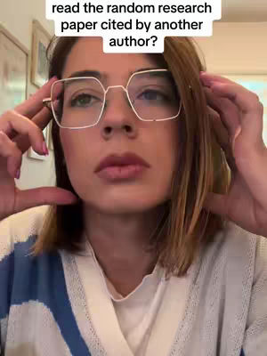 A close-up shot of a woman with light skin, wearing large square-framed glasses, gazing directly at the camera with a thoughtful expression. The background is slightly blurred and shows a glimpse of a room with what appears to be artwork on the wall. The woman is holding her hands up as if framing her face with a subtle gesture.