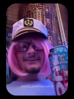 A person with a pink wig, glasses, and a sailor's hat is holding a can of soda. The background has some kind of a gold design on it. There is text saying 'HOPELESSSCOREHUB' across the bottom.