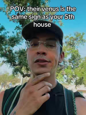 A selfie-style shot of a man wearing a black cap and glasses, with a blurred background of trees and a clear blue sky. The man is looking towards the camera with a thoughtful expression, hand touching his chin. Text overlay at the top reads: 'POV: their venus is the same sign as your 5th house'.