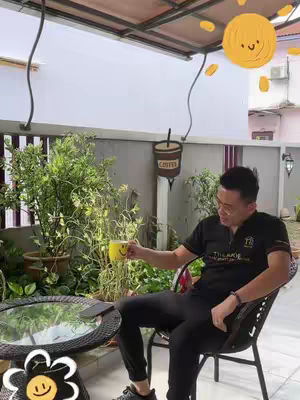 A man sits in an outdoor seating area, holding a yellow mug with a smiley face. The scene is well-lit, with lush greenery in the background and a table in front of him. The image has a casual, relaxed atmosphere, suggesting a peaceful setting.