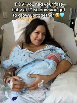 A woman lying in a hospital bed, holding a newborn baby. She is smiling and looking at the camera. The baby is wrapped in a blue blanket and wearing a pink hat. The background includes hospital equipment.