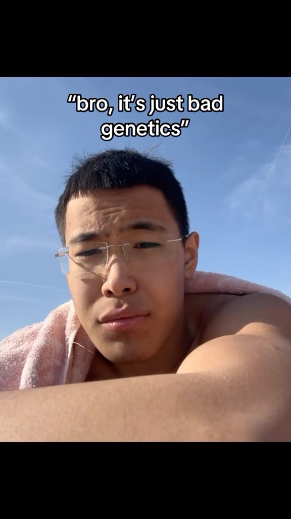 A selfie-style shot featuring a young man against a backdrop of a blue sky. He wears glasses and has a pink towel draped over his shoulders, suggesting a relaxed, outdoor setting. Text overlayed reads "bro, it's just bad genetics".