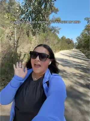 A woman in sunglasses waves at the camera while walking on a path with trees on either side. The sky is blue, and the light is bright. Text overlayed reads, 'I walk not to race anyone but to quiet my mind.'