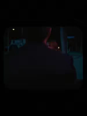 The image shows a dark, low-lit scene, viewed through what appears to be a frame or window. The primary subject is the back of a person, possibly a figure in silhouette, with their form filling a significant portion of the frame. Beyond this figure, glimpses of a city scene are barely visible, hinting at a nighttime environment with lights contributing to the dim ambience
