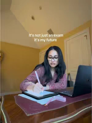 A woman with long dark hair and glasses sits at a desk in a room. She is wearing glasses and is writing on a tablet with a stylus. In front of her is a laptop, and behind her is a white door and a yellow painted wall.