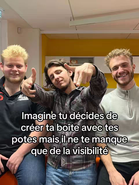 Three young men are posed in a medium shot, one in the center pointing towards the viewer with both hands, while the other two on either side smile and give a thumbs-up. The background suggests an office or classroom setting with a bright yellow wall.