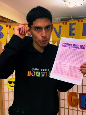 A young man in a black hoodie holds a pink paper with a crossword grid, pointing to his head. He's standing in front of a banner with colorful letters, with an emoji sticker and a decorative backdrop.