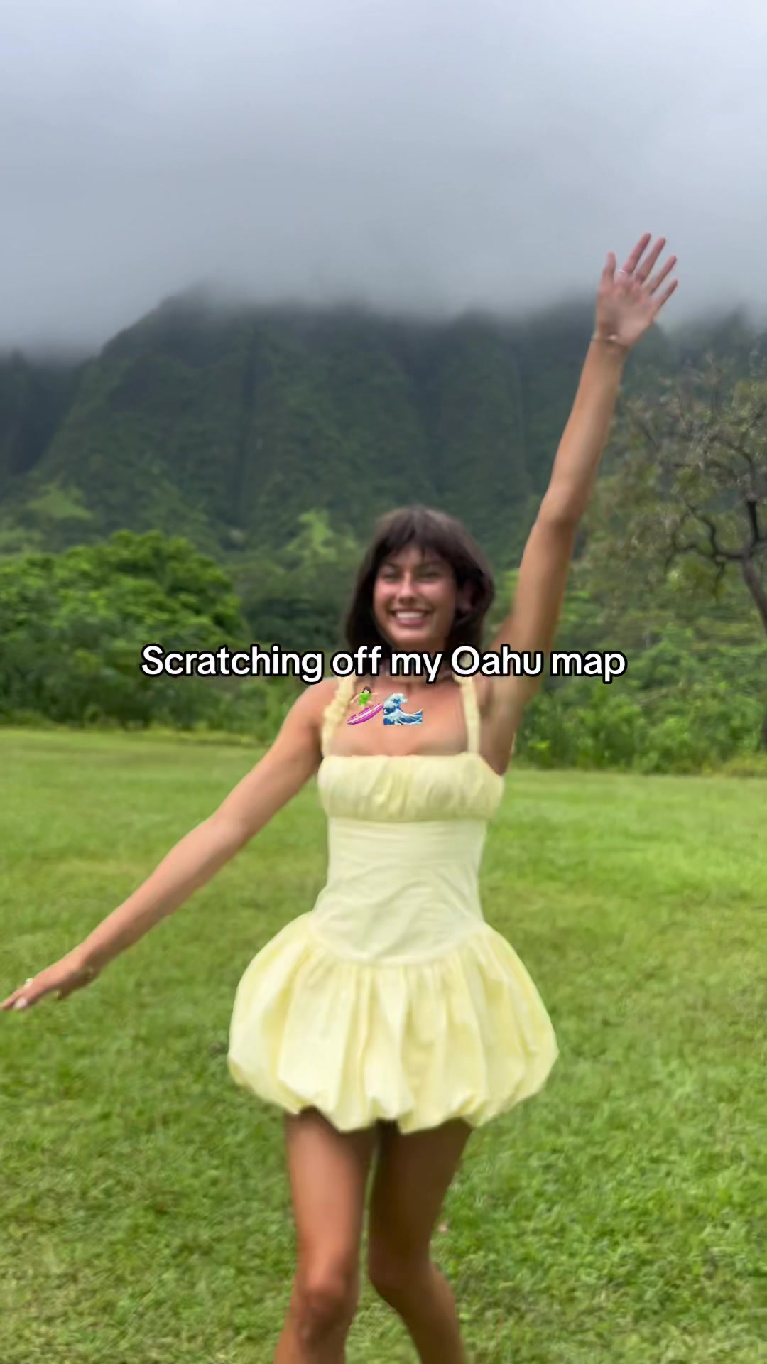A woman in a yellow dress stands in a grassy field with arms raised, smiling. Text overlays read "Scratching off my Oahu map". Behind her are green hills and a cloudy sky.