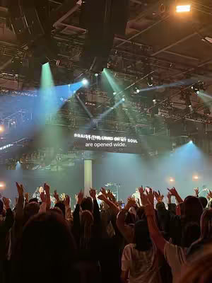 The image shows a large indoor space lit by spotlights and stage lights during a concert or worship service. The crowd has their arms raised, and there's a display with text on the back of the stage.