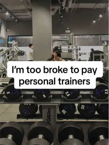 A selfie taken in a gym setting. The person is wearing a cap, holding a phone up to take the photo, and the frame shows the person in the gym surrounded by dumbbells and exercise machines. The background contains other people exercising.