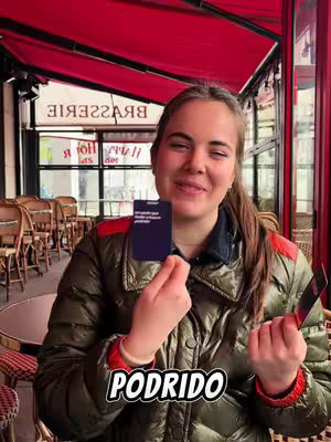 A woman is posing in front of a brasserie, holding two cards with a logo across the bottom saying 'PODRIDO'.