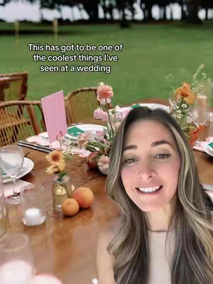 The cover image shows a light-skinned woman smiling at the camera, with her face filling most of the right side of the frame. Her long, light-brown hair has blond highlights framing her face. She wears a neutral colored top. The background is out of focus, but appears to be a wedding reception setting outdoors with a table setting in the foreground. The hook text is at the top and reads, "This has got to be one of the coolest things I've seen at a wedding."