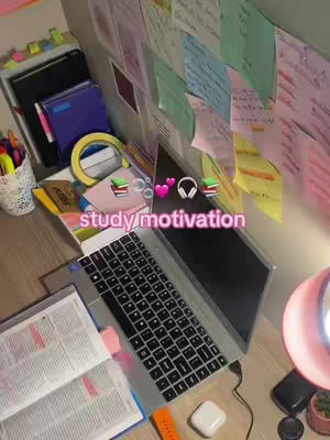 A top-down shot of a desk setup with a laptop open. A wall covered in sticky notes is visible in the background. The text overlay says “study motivation” in a pink, bubbly font.