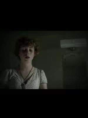 The cover image shows a dark, indoor scene. A young woman with brown, curly hair is the focus, positioned in the lower center. She wears a light-colored top with a floral pattern and a necklace. The lighting in the room is dim, making the background indistinct, with a small air conditioning unit visible to the right. The image has a grainy quality, enhancing a sense of mystery or suspense.