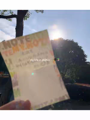 The image features a hand holding a slightly crumpled, light-colored sticky note against a sunny outdoor backdrop. The note has some text and a border, with a blank area suggesting it may be a to-do list or a note. The background includes a blurry view of a tree, the bright sun, and hazy sky. The overall effect is casual, suggesting a moment captured in everyday life.