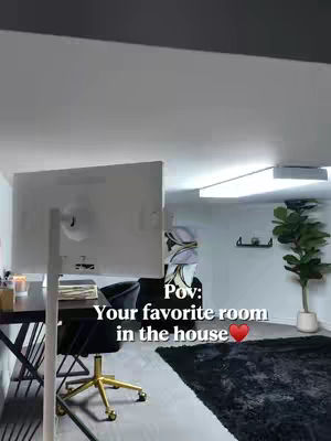An interior shot presents a modern, aesthetically pleasing room. A large computer monitor dominates the foreground, with a desk and chair positioned prominently. The background reveals a cozy lounge area with a black rug and a green plant, creating a balanced composition of work and leisure.