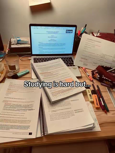 A top-down view of a wooden desk covered in study materials. A laptop is open in the back, displaying text on the screen. Notebooks and papers with highlighted text are spread around the laptop. A glass, pen holders with pens and highlighters are visible. Text overlayed on the image reads: Studying is hard but...