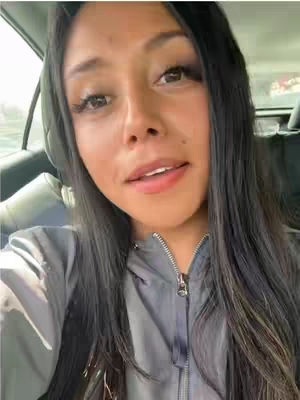 A woman is looking directly at the camera while sitting in a car. She has long, dark hair and is wearing a gray hooded sweatshirt. The background is blurred, suggesting movement.