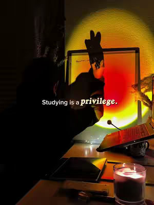 A dimly lit scene with a person's silhouetted figure in the foreground, partially obscuring a brightly lit area. The person's hand is raised towards a bright circle of orange and yellow light. A desk or table with various objects, including a candle, phone, and laptop, fills the lower part of the frame, adding to the study setup. The text "Studying is a privilege" is overlaid on the image.