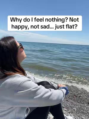 A woman is sitting on a rocky shore, facing away from the camera and looking upwards. She is wearing sunglasses, a light grey sweatshirt, and dark pants. The background features a vast ocean with a clear blue sky. A text overlay in a white box is positioned at the top of the image.