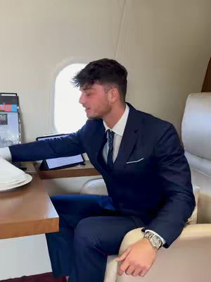 A man in a navy suit and tie sits at a table in a private jet cabin. The window is visible behind him along with the interior decor. The man is leaning, and there are sheets on a table and a screen in front of him