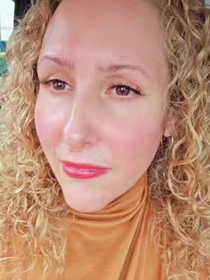 A close-up shot of a woman's face. She has curly blonde hair, fair skin, and is wearing a gold turtleneck. Her cheeks are flushed, and she has red lipstick on. The lighting seems soft and natural.