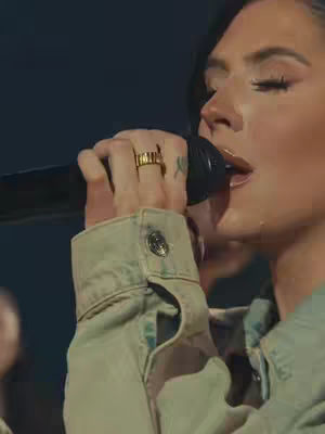 A medium shot features a woman close to a microphone, singing. The woman is light-skinned, with dark hair and wearing a light green jacket. A gold ring is on her finger. The background is a dark, indistinct space.