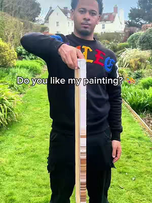 A man in a black sweater stands outdoors, holding a painting. The background includes trees, bushes, and a house.