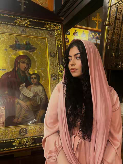 A young woman stands indoors in a church setting. She wears a pink headscarf that drapes around her face and shoulders, with long, dark curly hair visible. Behind her, an ornate gold-framed religious icon is visible. The image features a warm color palette, with the woman's attire and the icon's details creating a serene ambiance.