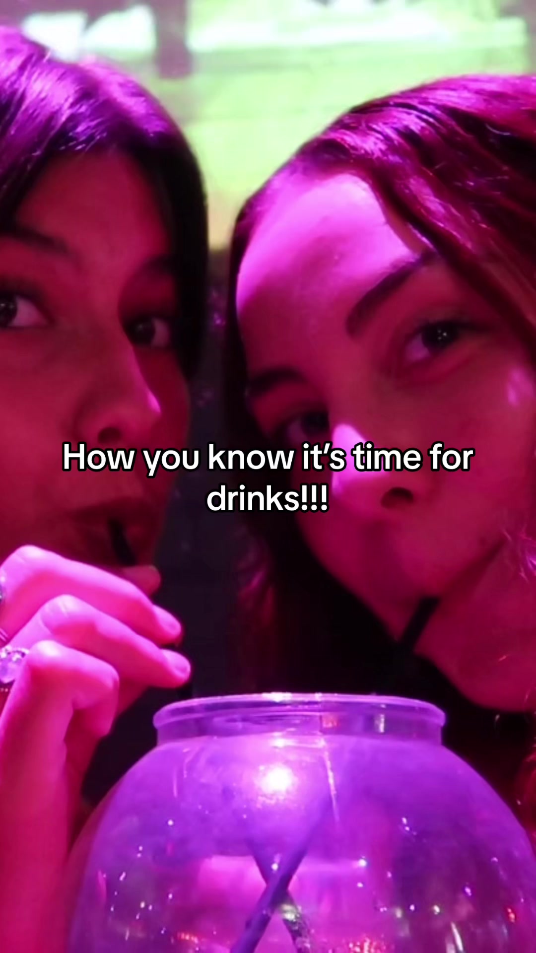 Two young women are shown closely, almost in a selfie-style angle, with a large, round glass container in the foreground. The background is illuminated with purple and green lights. Text overlayed reads, 'How you know it's time for drinks!!!'.