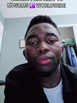 A man of African descent stares intently at the camera in a medium shot. The person is positioned in the center of the frame, wearing a black jacket over a white shirt. In the background, there is a light-colored wall, and a glimpse of a closet with clothes is visible on the right side. The top of the frame has overlaid text.