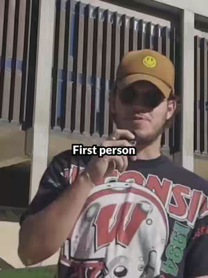 A man in a brown cap and a Wisconsin t-shirt looks directly at the camera. The background features a series of vertical metal slats.