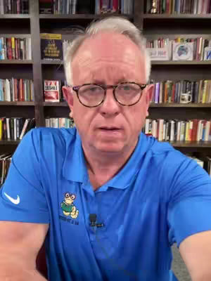 A man with glasses wearing a blue polo shirt sits in front of a bookshelf. The background is filled with books, creating a cozy and intellectual atmosphere. The man is centered in the frame and looks directly at the camera.