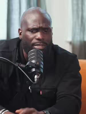 A bald, dark-skinned man with a beard is positioned in front of a microphone. He is wearing a black jacket. The background consists of a lightly colored curtain.