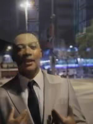 A man in a suit stands outside at night, with a city backdrop. The background is blurry with streetlights and illuminated buildings. The man is in what appears to be a beige suit with a black tie. He is gesturing with his hands as if he is talking.