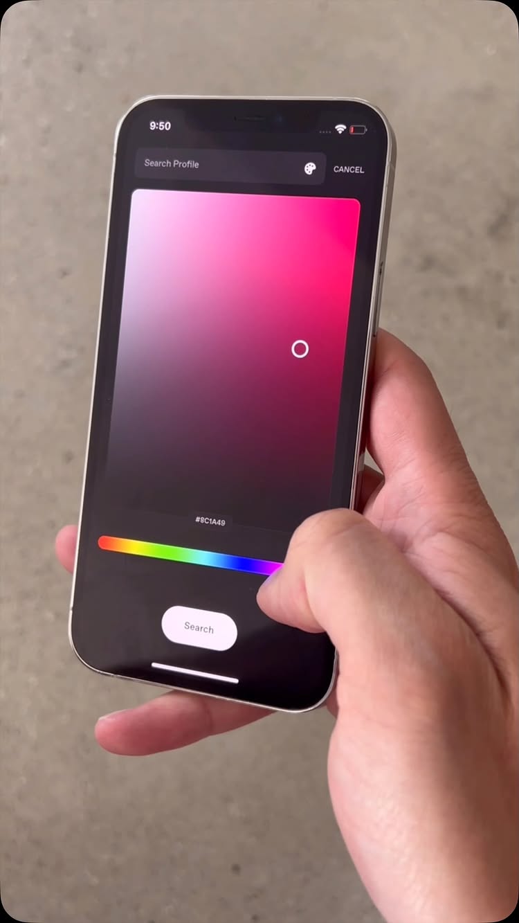 A hand holding a silver smartphone displaying a color gradient. The phone screen shows a gradient from red to pink, with a spectrum of colors at the bottom. A finger is touching the screen. The background is a grey stone surface.