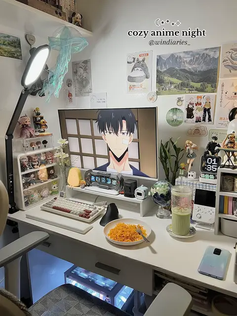 A well-lit desk setup with an anime theme featuring a computer monitor displaying anime characters, a plate of noodles, and various decorative items such as figurines and an aesthetic desk lamp. The composition suggests a cozy and organized workspace.