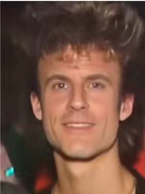 A close-up shot of a man's face, capturing his expression. He has short, dark hair with a curly texture on top. His eyes are looking towards the camera. The background is blurred and indistinct.
