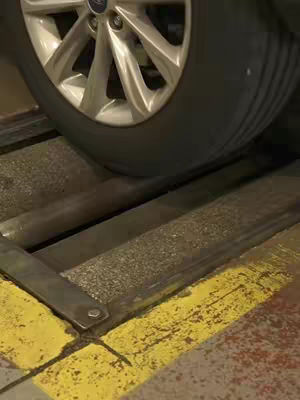 A close-up shot of a car tire resting on a car testing machine. The tire is black, set against a machine that has yellow borders. The background is a dimly lit garage type environment. The image is a static shot highlighting the vehicle's tire and the structure that evaluates it.