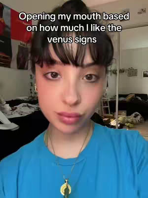A selfie of a woman with dark hair and bangs, looking at the camera. She has visible makeup, including blush and lip color. Behind her is a cluttered room with items like a bed and a red flag.